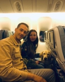 Ariella sitting on an airplane beside a man with their calm Golden Retriever service dog resting between them, demonstrating confident travel behaviour and companionship through Partner Dogs Canada.